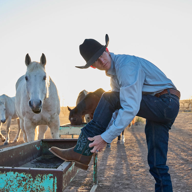 A man wearing a cowboy hat and bending down to put his boots on. 